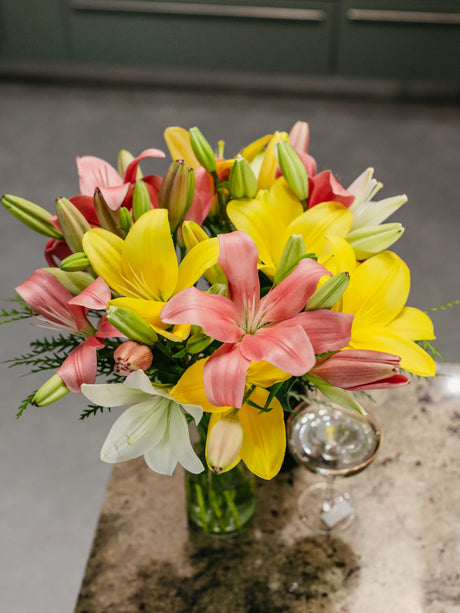 Bouquet of yellow, pink, and white lilies in a clear vase on a marble surface.