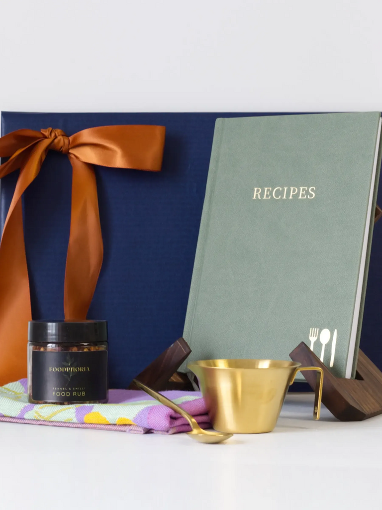 The primary item displayed is an A5 recipe keeper cookbook with a sage green linen cover and the word "RECIPES" embossed in gold. This book is resting on a minimalist, dark wooden stand.

In front, a gold-toned measuring cup and a matching small gold teaspoon sit on a colourful, patterned cotton kitchen towel. Next to the gold cup is a small jar of FOODPHORIA Fennel & Chilli Food rub. 