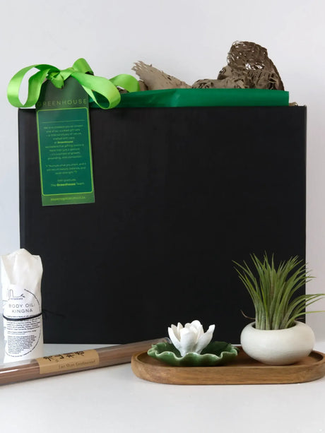 A gift set featuring a large black box tied with a green ribbon, a bottle of body oil, a bundle of incense sticks, a small white lotus flower in a green dish on a wooden tray, and an air plant in a white ceramic pot.