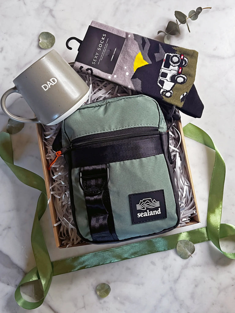 Father's Day gift box containing a green Sealand bag, silver "DAD" mug, and astronaut-themed socks, decorated with eucalyptus leaves and green ribbon on marble.