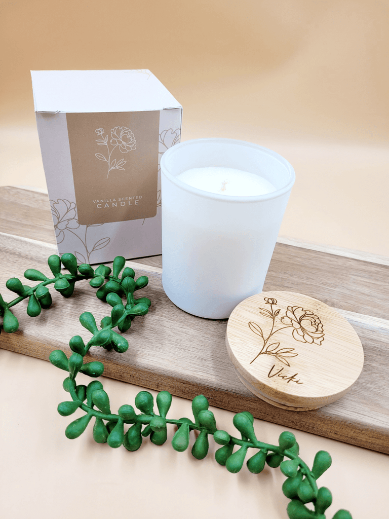 Personalised soy candle in white glass jar alongside engraved bamboo lid and floral printed packaging. 