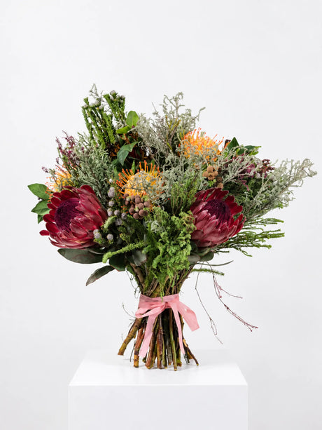 Tied bouquet of red proteas, orange pincushion proteas, brunia berries and fynbos foliage with a pink ribbon, on a white plinth