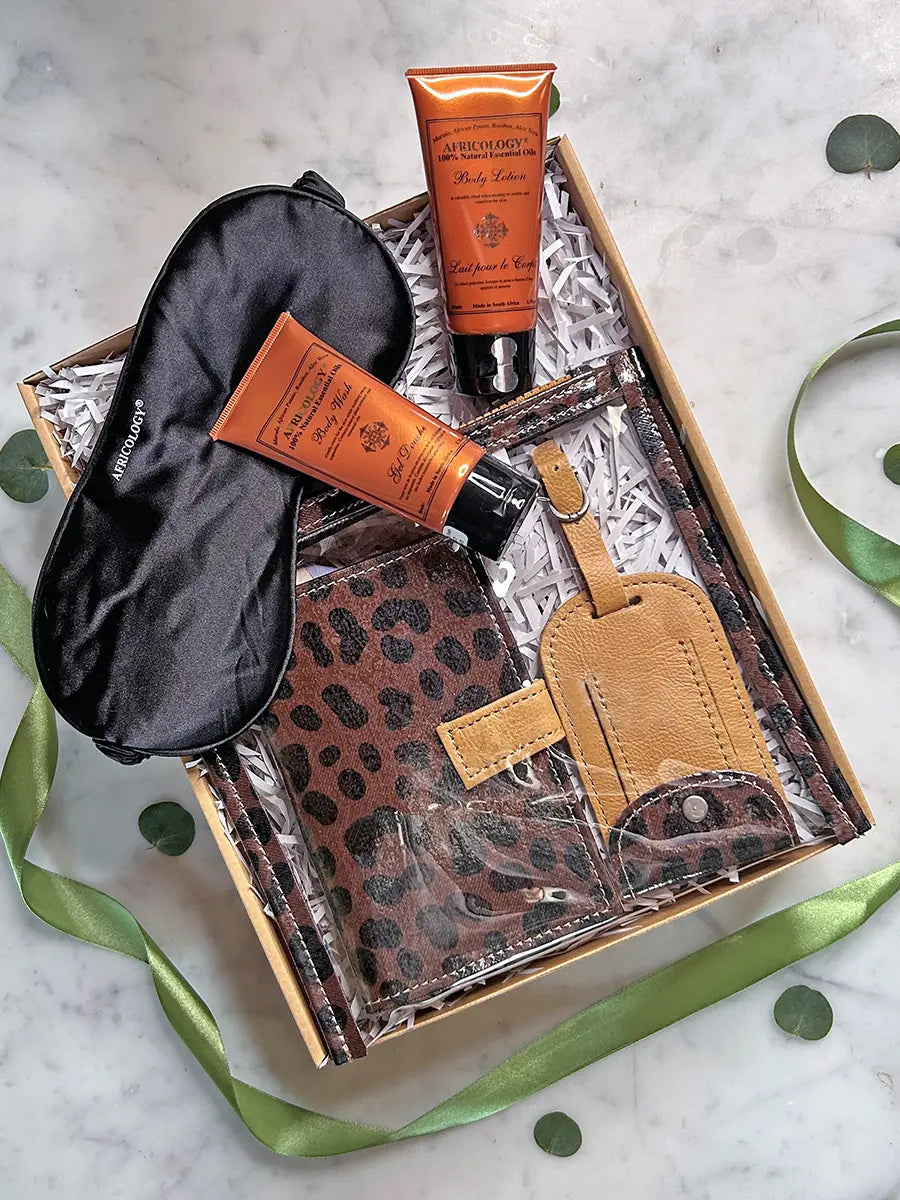 A curated gift set in a cardboard box with white crinkle paper, featuring a black satin sleep mask, two orange tubes from The Riches, a brown leopard-print pouch, a tan leather luggage tag, and a matching leather card holder, arranged on a marble surface with green ribbon and leaf accents.