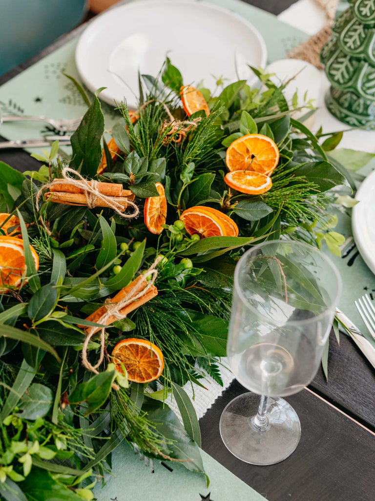 Rustic Christmas table centrepiece garland featuring dried orange slices, cinnamon sticks, pine, eucalyptus and mixed greenery styled on dinner table - natural holiday table runner for entertaining