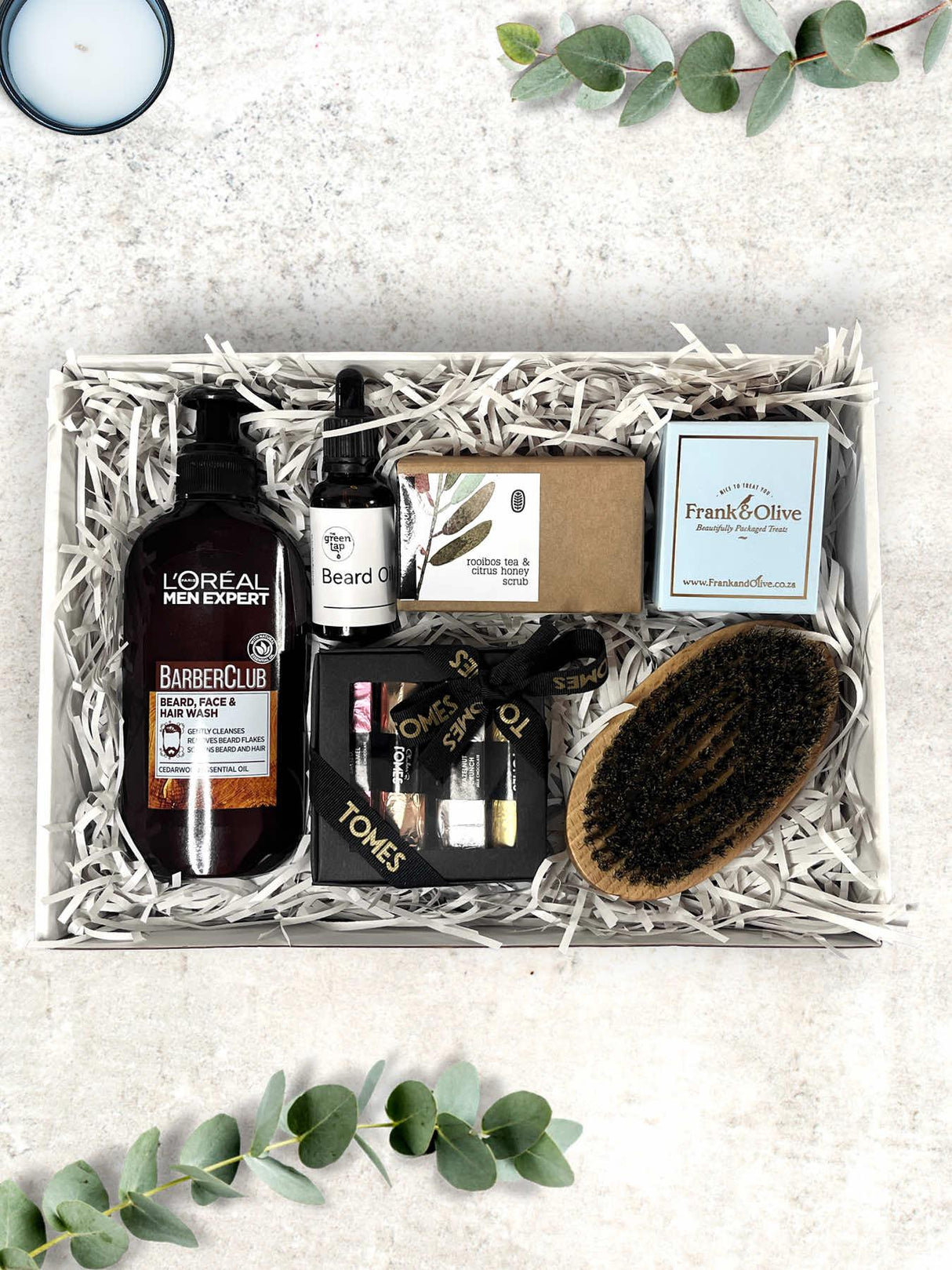 A gift box containing grooming products for men, including a beard brush, beard oil, beard and body wash, olive oil soap, chocolates, and fudge twisties.