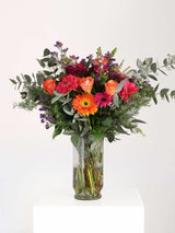 Bloomable's Celebration Bouquet displayed in a clear glass vase on a white pedestal. Features vibrant orange roses, pink and orange gerbera daisies, purple statice, and eucalyptus. Fresh flower arrangement perfect for special occasions with same-day delivery across South Africa.