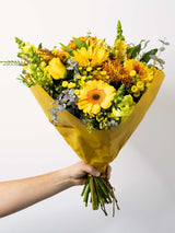 Bloomable's Happy Bouquet wrapped in yellow paper, held by a hand. Features bright sunflowers, yellow gerbera daisies, golden chrysanthemums, yellow roses, snapdragons, and eucalyptus accents. A cheerful arrangement available for same-day delivery across South Africa.