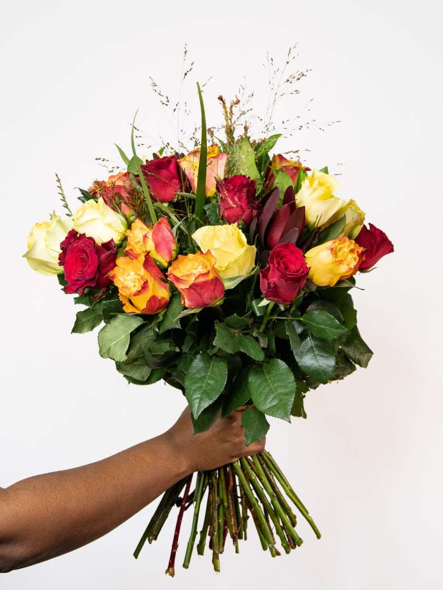 Bloomable's Mixed Roses bouquet hand-held against white background. Features a vibrant combination of deep red roses, sunny yellow roses, and bicolour orange-yellow roses with decorative grasses and fresh greenery. Perfect for Valentine's Day, Mother's Day, anniversaries, or expressing warm emotions with same-day delivery across South Africa.