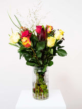 Bloomable's Mixed Roses bouquet in a ribbed glass vase on a white pedestal. Features red roses, yellow roses, and sunset orange-yellow bicolour roses with decorative grasses and rich green foliage. Perfect for Mother's Day, Valentine's Day, anniversaries, or expressing warm emotions with same-day delivery across South Africa.