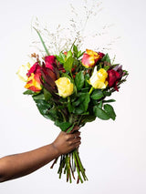 Bloomable's Mixed Roses bouquet held by a hand with ring. Features a beautiful arrangement of deep red roses, bright yellow roses, and sunset orange-yellow bicolour roses with decorative grasses and fresh greenery. Perfect for Valentine's Day, Mother's Day, birthdays, or expressing love with same-day delivery across South Africa.