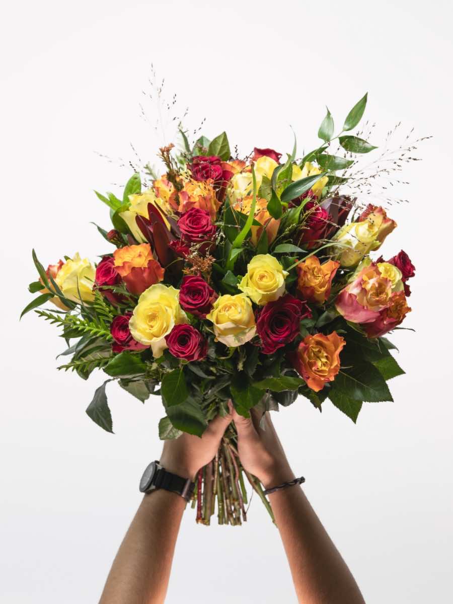Bloomable's Mixed Roses large bouquet held with both hands, featuring a luxurious arrangement of deep red, bright yellow, and sunset orange roses accented with fresh greenery and decorative grasses. The hands holding the bouquet display a watch and bracelet. Perfect for Valentine's Day, Mother's Day, anniversaries, or making a bold statement with same-day delivery across South Africa.