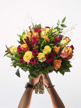 Bloomable's Mixed Roses large bouquet held with both hands, featuring a luxurious arrangement of deep red, bright yellow, and sunset orange roses accented with fresh greenery and decorative grasses. The hands holding the bouquet display a watch and bracelet. Perfect for Valentine's Day, Mother's Day, anniversaries, or making a bold statement with same-day delivery across South Africa.
