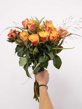 A hand holding an orange rose bouquet with lush greenery, created by South African florists, showcasing seasonal floral beauty.