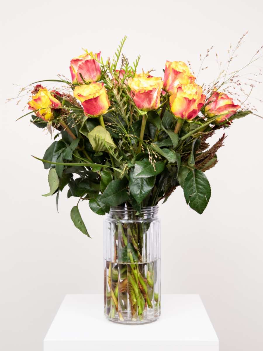 A vibrant orange rose bouquet with greenery, arranged by South African florists, displayed in a clear vase on a white pedestal.