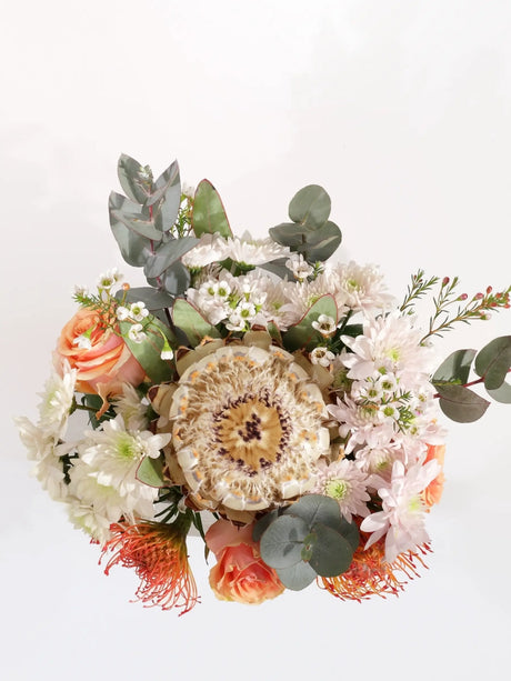 Overhead view of native flower arrangement with white protea, coral roses, orange pincushions, white chrysanthemums and eucalyptus in square glass vase - compact desk bouquet