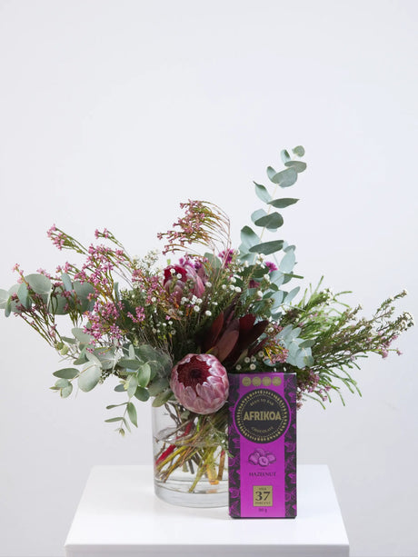 Bouquet of flowers in a glass vase with a box of chocolates on a white surface