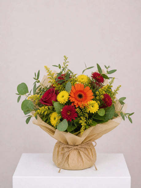 Bouquet of yellow, orange and red flowers with greenery wrapped in kraft paper.
