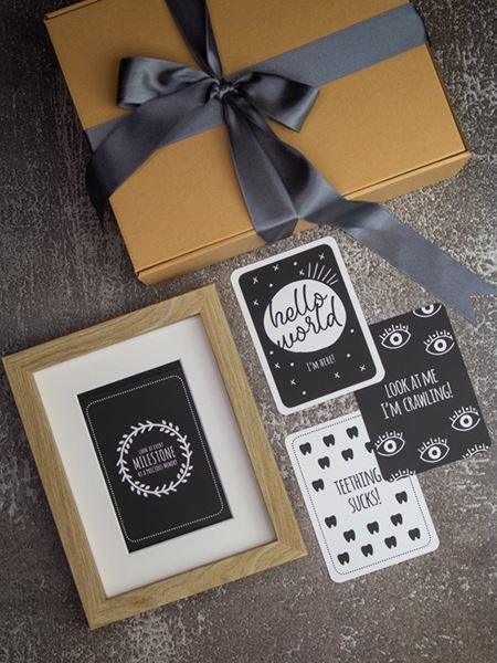 Milestone cards and wooden photo frame next to a gift box with ribbon, ideal for capturing and framing baby's first-year moments.