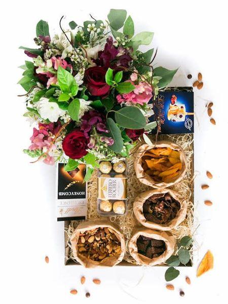 Decadent snack box with biltong, chocolate, nuts, and a vibrant floral arrangement in red, white, and pink hues.