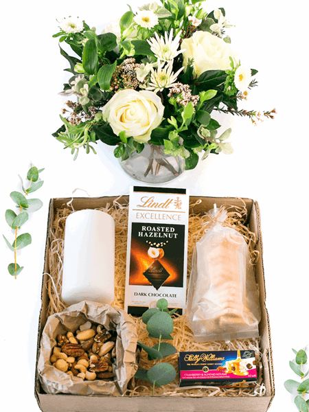 Serene gift box with white pillar candle, fresh floral arrangement, Lindt chocolate, luxury nougat, shortbread biscuits, and mixed nuts.