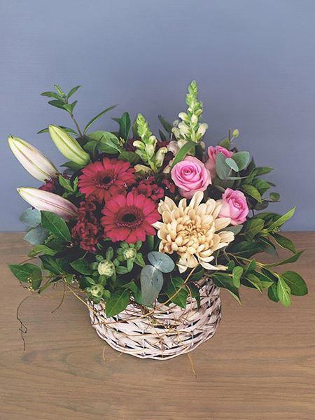 Colorful floral arrangement with cerise gerberas, pink roses, lilies, snapdragons, and peach chrysanthemum in a woven basket.