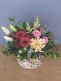 Colorful floral arrangement with cerise gerberas, pink roses, lilies, snapdragons, and peach chrysanthemum in a woven basket.