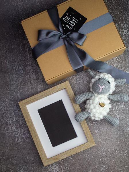 Gift box with 'Oh Baby' sticker, wooden photo frame, and crochet lamb toy on gray surface, tied with black ribbon.